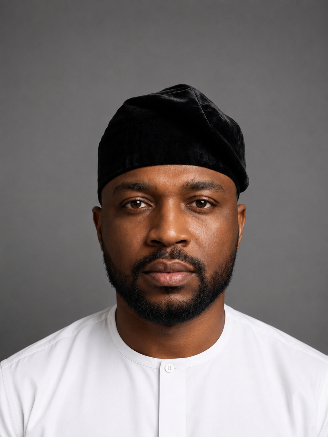 A man wearing a black velvet traditional cap and a white button-up shirt, photographed against a solid gray studio background.